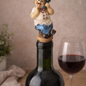 Puede incluir: Tapón decorativo para botella de vino con una figura de chef. El chef lleva un gorro blanco, una camisa blanca, un delantal azul y zapatos marrones. En el fondo se ve una copa de vino tinto.