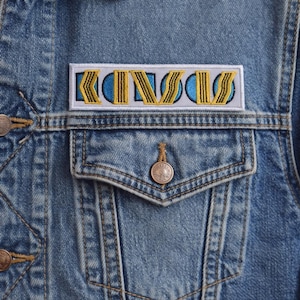 Peut inclure: Veste en jean bleu clair avec un écusson rectangulaire sur la poche poitrine. L'écusson affiche le mot "KINGS" en jaune, souligné de bleu et de blanc. La veste a une fermeture boutonnée et deux poches poitrine.