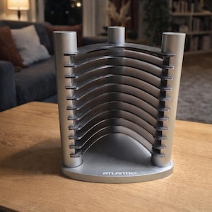 May include: A silver-colored CD or DVD storage rack with a curved design. The rack has a base with the brand name "ATLANTIC" printed on it. The rack is on a wooden table in a living room setting.