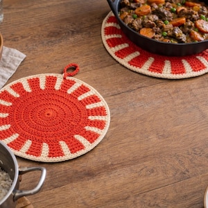 Puede incluir: Dos salvamanteles redondos de ganchillo, rojo y crema. Uno está en primer plano y el otro en segundo plano. Una sartén de hierro fundido con comida descansa sobre uno de los salvamanteles.