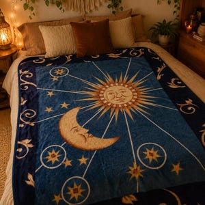Könnte beinhalten: Eine blaue Plüschdecke mit einer großen zentralen Sonne mit menschlichem Gesicht, einem Halbmond mit menschlichem Gesicht und verschiedenen goldenen Sternen, die durch feine Linien verbunden sind. Der dunkelblaue Rand zeigt ein florales Muster.