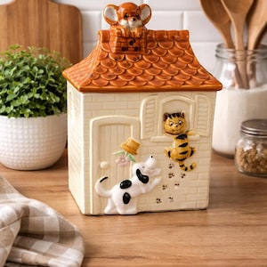 Peut inclure: Une boîte à biscuits en céramique en forme de maison avec un toit brun et des murs crème. Le pot représente une souris sur la cheminée, un chat à la fenêtre et un chien sur le côté. Le pot est décoré d'empreintes de pattes et d'un petit chapeau.