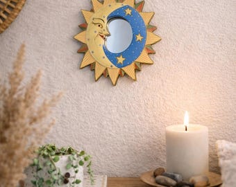May include: A celestial-themed wall mirror featuring a sunburst design with a smiling moon and stars. The mirror has a blue and yellow color scheme. The image also shows a lit candle, a small plant, and a wooden shelf.