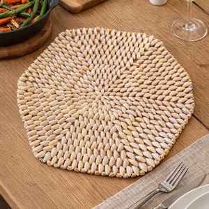 May include: Hexagonal beige placemat made of small, oval-shaped wooden beads. The placemat is on a wooden table, with a fork, knife, and plate visible. A pan of vegetables and a glass of wine are also in the image.