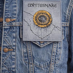 Peut inclure: Veste en jean bleue avec un écusson portant le nom du groupe "Whitesnake". L'écusson présente un motif circulaire avec des détails dorés et noirs. La veste a des boutons couleur laiton et des coutures en denim classiques.