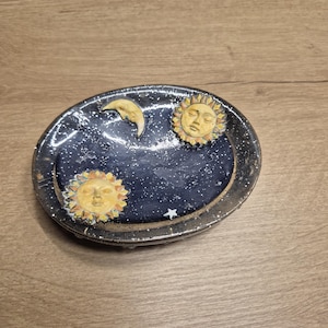 May include: An oval ceramic trinket dish with a celestial design. The dark blue interior is speckled with white dots, mimicking a starry night. It features two yellow suns and a crescent moon. The dish has a dark rim, creating a contrasting border.