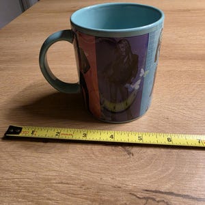 May include: A teal ceramic mug with a wraparound design featuring a person and butterflies. The mug has a teal interior and handle. A measuring tape is next to the mug, showing a length of approximately 6 inches.
