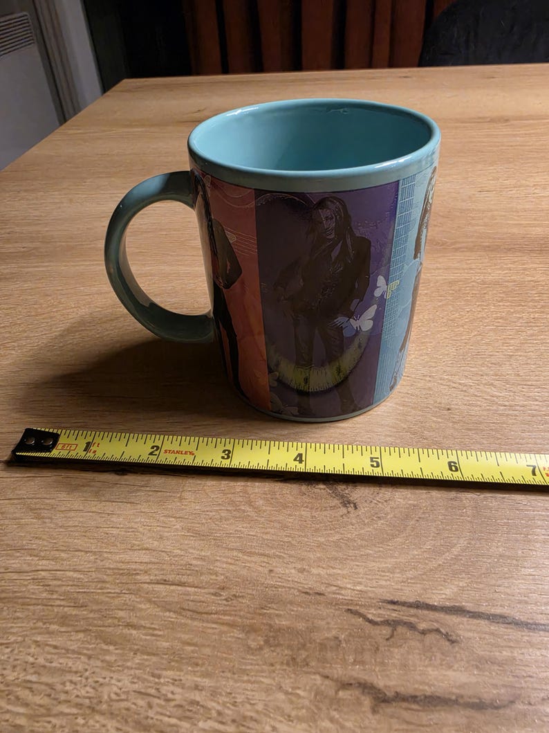 May include: A teal ceramic mug with a wraparound design featuring a person and butterflies. The mug has a teal interior and handle. A measuring tape is next to the mug, showing a length of approximately 6 inches.