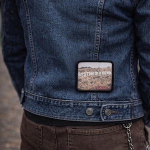 Peut inclure: Veste en jean bleu foncé avec un écusson portant les mots "SYSTEM OF A DOWN" et "TOXICITY". L'écusson présente un paysage désertique. La personne porte un pantalon marron.