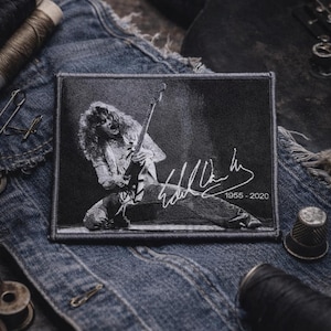 Peut inclure: Écusson noir et blanc représentant un musicien jouant de la guitare, avec une signature et les dates 1955-2020. L'écusson rectangulaire est bordé de coutures et posé sur une veste en jean.