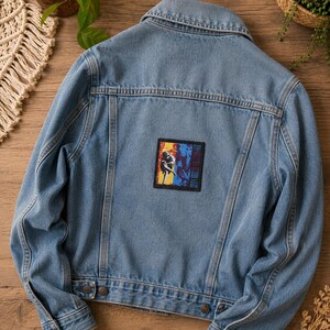Op de afbeelding: Een lichtblauw spijkerjack met een omgeslagen kraag, lange mouwen met knoopmanchetten en een rechthoekige patch op de rug. De patch toont een kleurrijke afbeelding met de tekst Guns N' Roses in een verticale oriëntatie.
