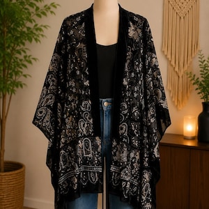 Op de afbeelding: Een zwarte fluwelen kimono met een transparante mesh-laag met een ingewikkeld paisley- en bloemenpatroon in goud- en crèmetinten. Het kledingstuk heeft een effen zwarte fluwelen bies langs de opening aan de voorzijde.