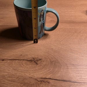 May include: A teal ceramic mug with a wraparound graphic of a cartoon character. The mug is approximately 4 inches tall. The handle is a matching teal color. The interior of the mug is also teal.