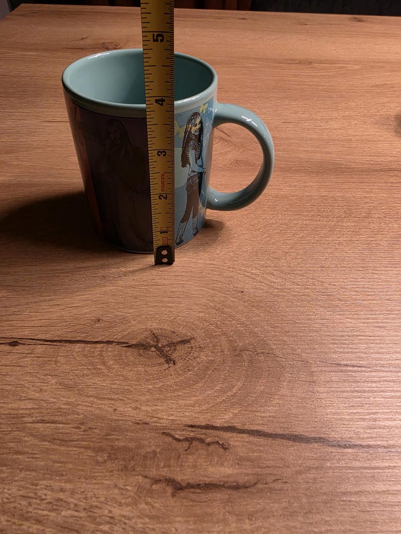 May include: A teal ceramic mug with a wraparound graphic of a cartoon character. The mug is approximately 4 inches tall. The handle is a matching teal color. The interior of the mug is also teal.