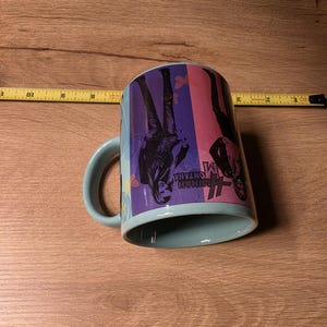 May include: A light blue ceramic mug with a curved handle. The mug features a colorful design with the words "Hannah Montana" and images of a person. The mug is resting on a wooden surface.