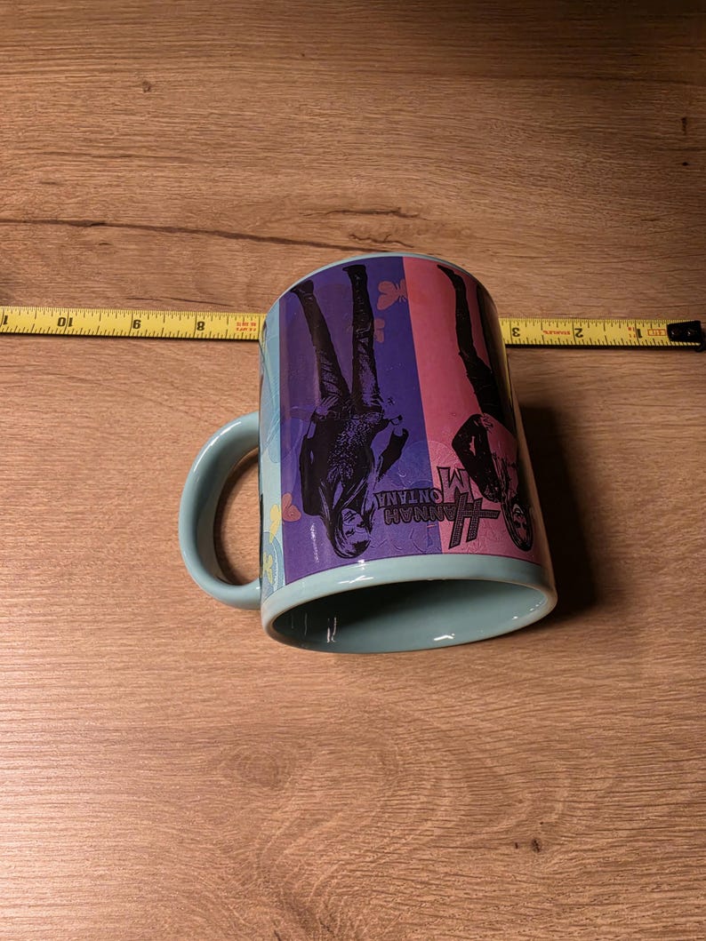 May include: A light blue ceramic mug with a curved handle. The mug features a colorful design with the words "Hannah Montana" and images of a person. The mug is resting on a wooden surface.