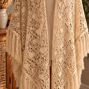 May include: A cream-colored crochet shawl featuring an openwork grid pattern interspersed with small heart motifs. The garment has long, straight fringe detailing along the bottom edges and is displayed on a mannequin torso.