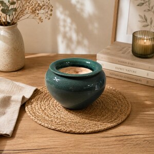 Peut inclure: Un pot en céramique vert d'eau à surface texturée est posé sur un dessous de plat rond en jute tressé. Un vase avec des fleurs séchées et une bougie allumée sont en arrière-plan. Le pot est sur une table en bois.