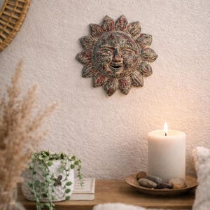 May include: A rustic sun-shaped wall hanging with a smiling face, painted in shades of red, brown, and gray, is mounted on a textured white wall. A lit white candle sits on a wooden surface, alongside a small plant and decorative stones.