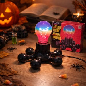 Peut inclure: Une lampe Lite Bulb avec une ampoule aux couleurs de l'arc-en-ciel, posée sur une base noire avec des pieds arrondis. La boîte à côté porte la même image. Thème Halloween avec citrouille, araignées et bonbons.