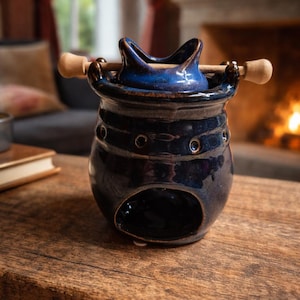 Puede incluir: Un difusor de aceites esenciales de cerámica azul oscuro con un asa de madera. El difusor tiene un cuerpo redondo con una gran abertura y varios agujeros más pequeños. La parte superior es azul claro con un diseño curvo. Está sobre una mesa de madera.