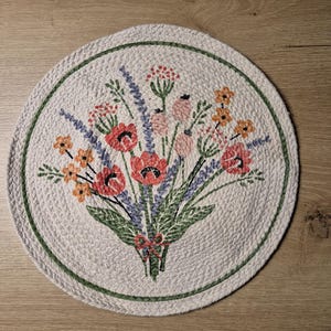 Puede incluir: Posavasos redondo, blanquecino, con una textura trenzada y un borde verde. El centro presenta un ramo de flores bordado en rojo, naranja, rosa y morado. Un elemento decorativo para la mesa.