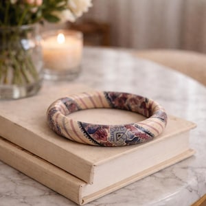 May include: A patterned bangle bracelet with a cream base and a design in red, blue, and purple. The bracelet sits on two stacked books. A lit candle and flowers are in the background. The bracelet's diameter is approximately 7 cm.