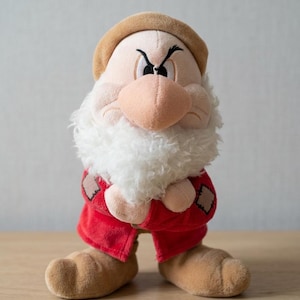 May include: A plush Grumpy doll from Snow White and the Seven Dwarfs. The doll features a beige hat, nose, and feet, a white beard, and a red outfit with brown patches. The doll has an angry expression.