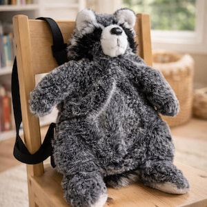 May include: A grey and white plush raccoon backpack with black straps sits on a wooden chair. The backpack has a white face and black eyes. The chair is light brown and the background is blurred.