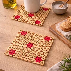 Puede incluir: Dos posavasos cuadrados de madera con diseño floral y detalles rojos. Un posavasos sostiene una taza de café blanca. Los posavasos están sobre una mesa de madera marrón claro, con otros objetos en el fondo.