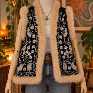 Könnte beinhalten: Eine schwarze ärmellose Weste mit weißer Blumenstickerei und einem dicken beigen Kunstfellbesatz an Kragen, Armausschnitten und Saum. Die Weste wird über einem weißen Oberteil und einer blauen Jeans getragen und mit einer runden Metallkette ergänzt.