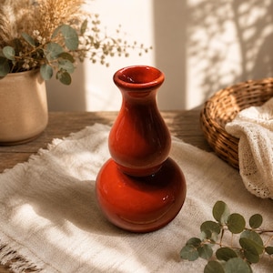 Peut inclure: Un vase en céramique rouge brillant, avec une base arrondie et un col étroit. Le vase est posé sur un tissu en lin beige. Un panier en osier et des fleurs séchées sont en arrière-plan. L'éclairage crée des ombres.