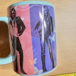 May include: A ceramic mug with a light blue handle and rim. The mug features a split design with a woman in black pants and a jacket on a pink background and a purple background. Butterflies are also visible.