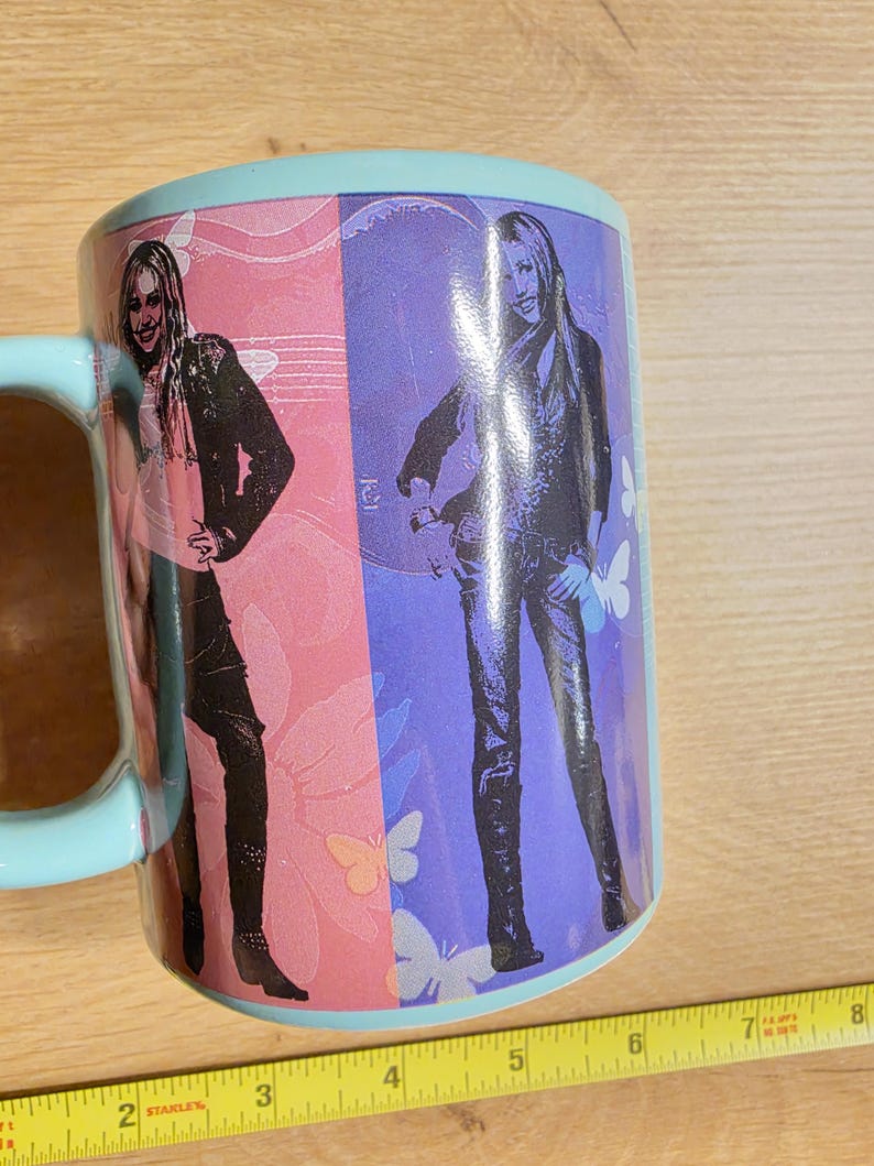 May include: A ceramic mug with a light blue handle and rim. The mug features a split design with a woman in black pants and a jacket on a pink background and a purple background. Butterflies are also visible.