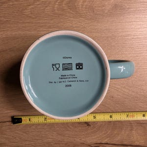 May include: A light blue ceramic mug with a matching handle. The bottom of the mug has the Disney logo and text that reads "Made in China" and "2008". A yellow measuring tape is next to the mug.