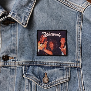 Peut inclure: Une veste en jean bleu clair avec un écusson rectangulaire brodé sur la poitrine. L'écusson présente une photographie du groupe Whitesnake avec le nom du groupe écrit en lettres blanches au-dessus des membres jouant des instruments.