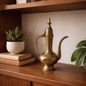May include: A vintage brass pitcher with an ornate design, featuring a long spout and a curved handle. The pitcher is displayed on a wooden shelf alongside a potted succulent and a stack of books, creating a decorative display.