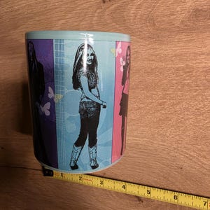 May include: A ceramic mug with a light blue rim and a colorful design featuring three images of a woman. The mug has a light blue background with butterflies and musical notes.