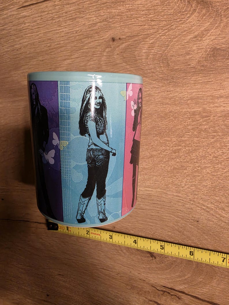 May include: A ceramic mug with a light blue rim and a colorful design featuring three images of a woman. The mug has a light blue background with butterflies and musical notes.