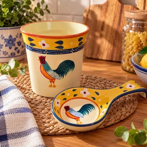 Peut inclure: Un pot à ustensiles et un repose-cuillère en céramique assortis, présentant une base couleur crème avec un motif de coq peint à la main, des bords jaunes ornés de fleurs blanches et des lignes décoratives bleu foncé.