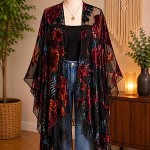 May include: A sheer black kimono featuring a velvet floral pattern in shades of deep red, orange, and teal. The garment has an open front, wide flowing sleeves, and a handkerchief hemline, layered over a black top and blue denim jeans.