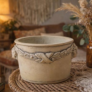 Peut inclure: Un pot de fleurs rond beige avec un motif décoratif de couronne de laurier. Le pot a deux petites poignées et repose sur une surface en osier tressé. Un vase de fleurs séchées est en arrière-plan.