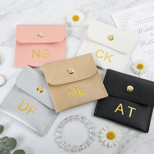 May include: A collection of personalized jewelry pouches in pink, white, gray, tan, and black, each with gold initials. A wedding invitation and jewelry, including rings and a bracelet, are also present, along with daisies.