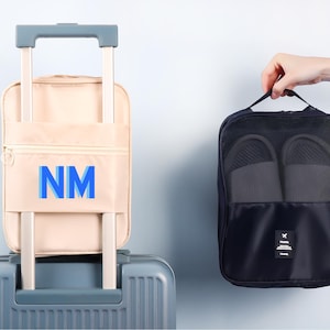 Puede incluir: Una maleta beige con las letras azules "NM" en la parte delantera y una bolsa de zapatos azul marino con dos pares de zapatillas grises. La bolsa de zapatos está sujeta por una mano.