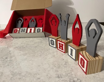 May include: Wooden block letters spelling "OHIO" with red and gray figures. The figures are in various poses, some with arms raised. The blocks are in a cardboard box with red lining and also arranged separately.