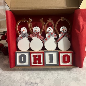 May include: A decorative Ohio-themed Christmas ornament set. Four snowman figures with wooden antlers and scarves are displayed above blocks spelling "OHIO". The snowmen are in a red box with red tissue paper.