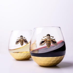 May include: Two stemless wine glasses with gold honeycomb bases and bee embellishments. One glass contains white wine, the other red wine. The bees are detailed with black and clear crystals. The glasses are on a white background.