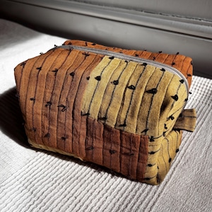 May include: A rectangular fabric pouch featuring a patchwork design of mustard yellow and burnt orange panels. The exterior is quilted with vertical stitching and accented by small, dark black thread knots distributed across the surface.