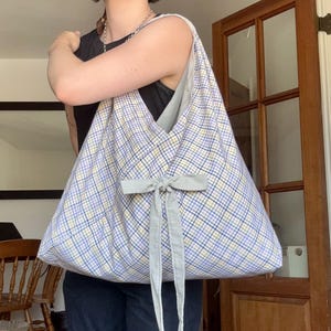 以下が含まれることがあります： 青、黄、白のチェック柄の生地で作られた大きなホーボータイプのショルダーバッグ。バッグは柔らかく型崩れしやすい形状で、前面中央にライトグレーの布製リボン結びがあり、2本の長いリボンの端が垂れ下がっています。