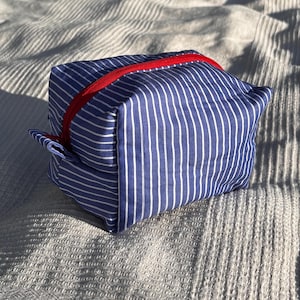 Può includere: Una pochette rettangolare in tessuto con un motivo a righe verticali sottili blu e bianche. La borsa è dotata di una chiusura a cerniera rosso brillante che corre lungo la parte superiore e di una piccola linguetta laterale in tessuto.